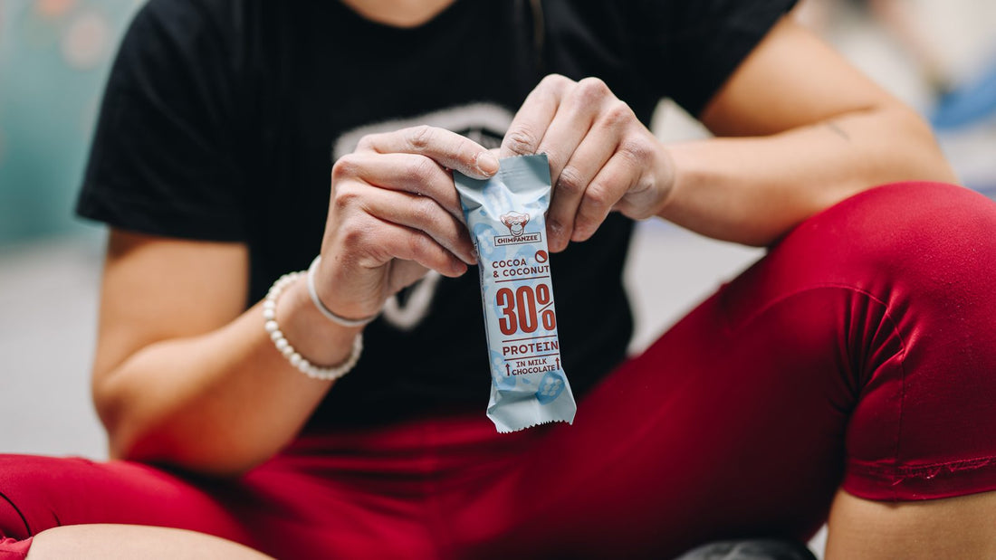 Athlete in a black T-shirt and red leggings sitting on an indoor gym floor, opening a Chimpanzee 30% Protein Bar Cocoa & Coconut